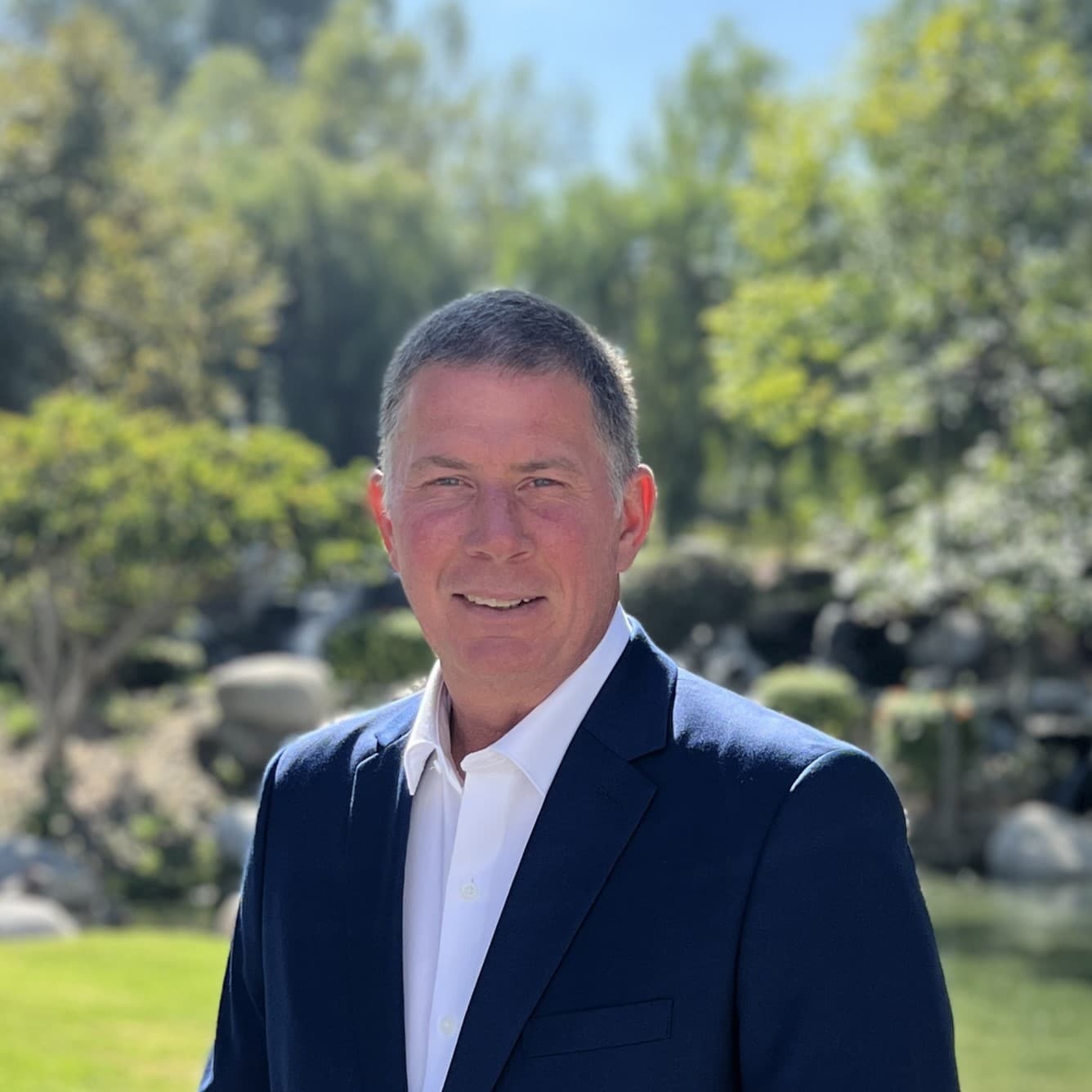 Professional headshot of a man in a navy suit standing outdoors in a landscaped garden setting with trees and sunlight in the background.